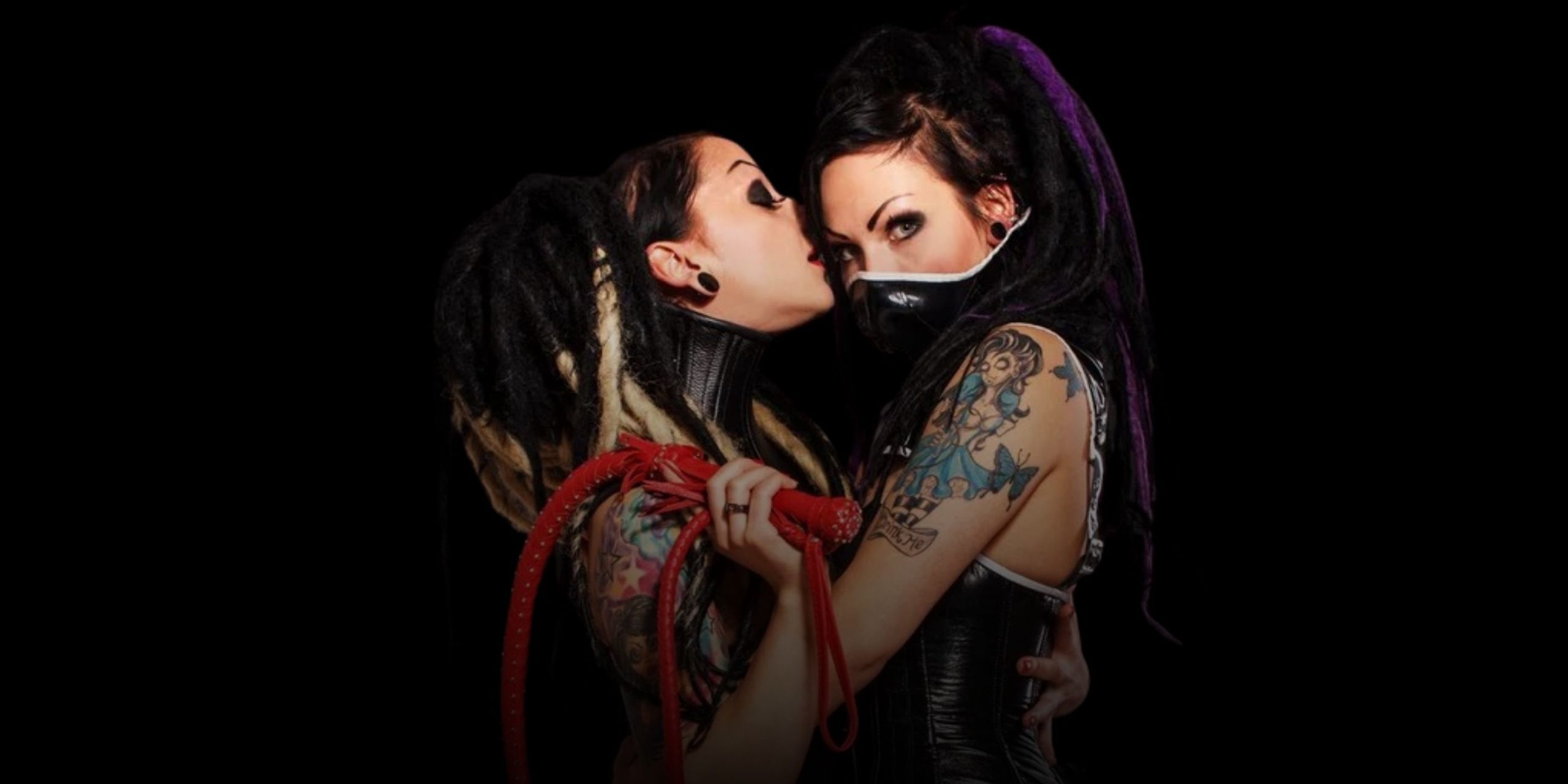 Two goth looking women embrace while one holds a red whip.